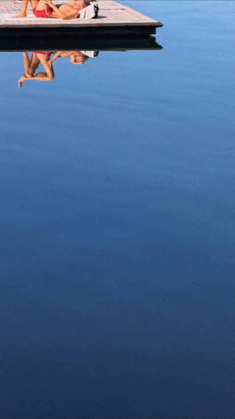 Un pontile sull'acqua blu immobile, con una persona sdraiata riflessa a testa in giù